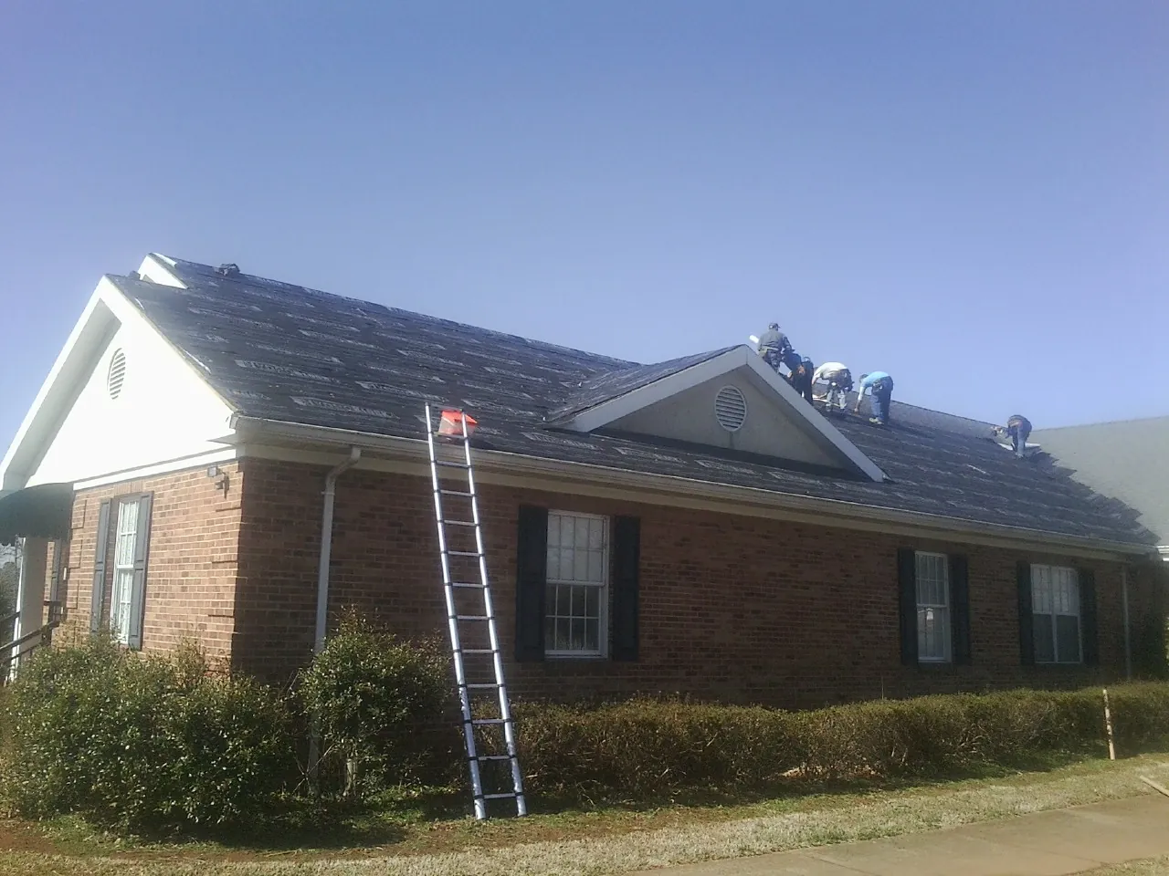 Crew tearing shingles off a brick ranch roof with a ladder set — Emergency Roof Repair work by our crew in Constantia